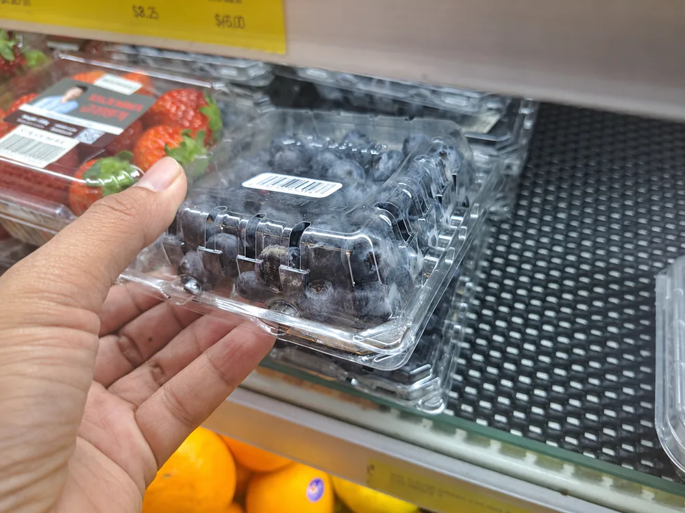 Shopper hand selecting a clear PET blueberry clamshell from a refrigerated retail shelf showing consumer choice
